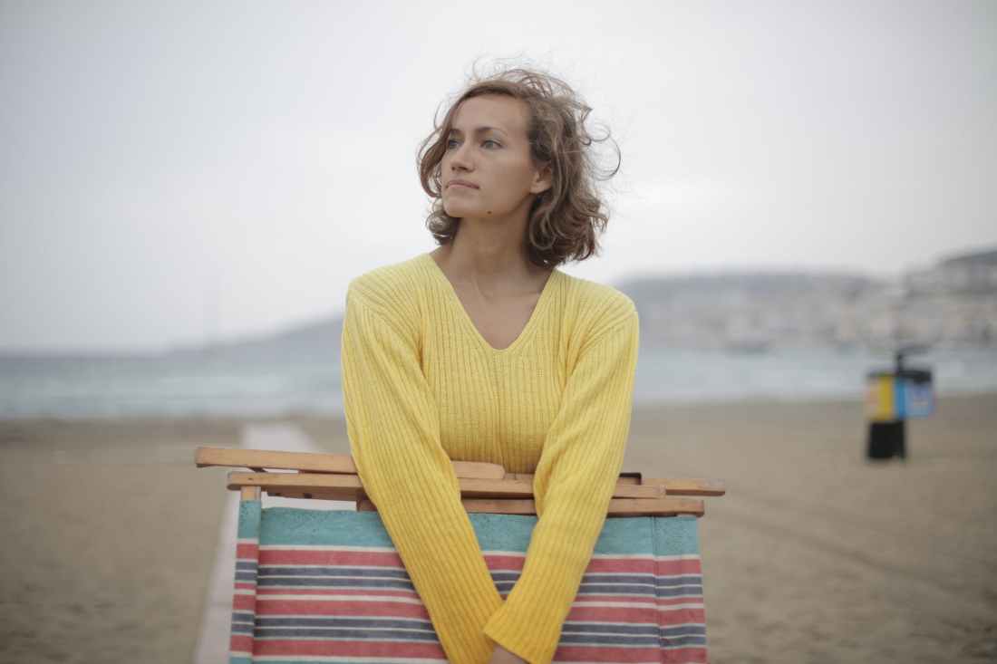 calm female tourist with folded deckchair standing alone on seashore in overcast weather